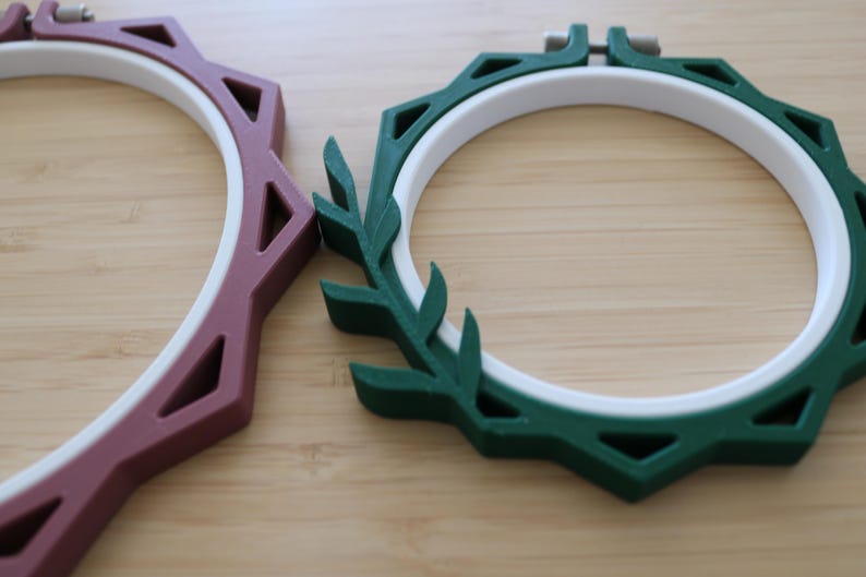 May include: Two circular embroidery hoops featuring white inner rings and colored outer frames. One frame is dark green with a leaf-like design on one side, and the other is a muted mauve with a geometric, angular, and faceted outer edge.