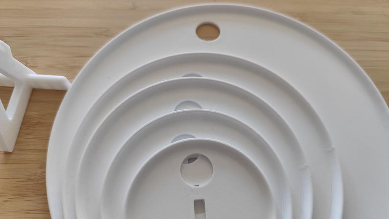 May include: A set of four nested white circular plastic discs of varying sizes, each featuring a centered circular hole and a small rectangular cutout at the bottom. A white plastic stand is positioned to the left of the stacked discs.