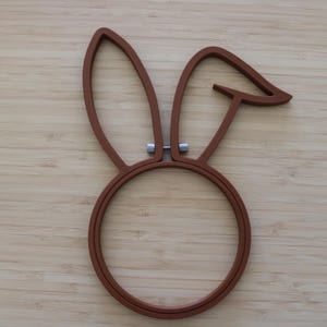 May include: A brown embroidery hoop shaped like a rabbit head with two long ears, one of which is bent at the top. The hoop features a circular base and a small silver-colored metal tightening screw located between the two ears at the top.