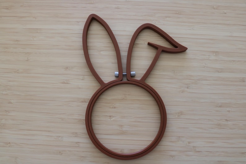 May include: A brown embroidery hoop shaped like a rabbit head with two long ears, one of which is bent at the top. The hoop features a circular base and a small silver-colored metal tightening screw located between the two ears at the top.