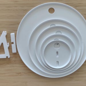 May include: A set of five white circular plastic plates of varying sizes nested together, each featuring a small circular hole near the top edge. Beside the nested plates is a white triangular plastic stand with a separate rectangular connector piece.