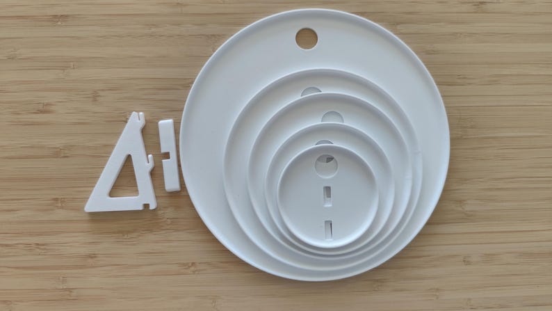 May include: A set of five white circular plastic plates of varying sizes nested together, each featuring a small circular hole near the top edge. Beside the nested plates is a white triangular plastic stand with a separate rectangular connector piece.