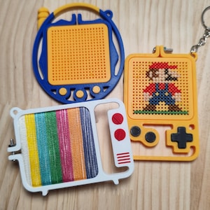 May include: Three plastic embroidery hoops shaped like retro electronics. One is a blue circular hoop with a yellow grid, one is a yellow handheld game console with a cross-stitch Mario character, and one is a white television frame with colorful thread.