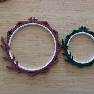 May include: Two circular embroidery hoops with white inner rings and colored outer frames. The larger hoop is maroon with a leaf-shaped extension on the left side. The smaller hoop is dark green with a matching leaf-shaped extension on the left side.