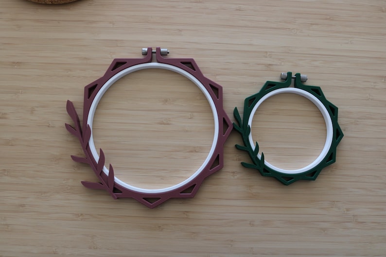 May include: Two circular embroidery hoops with white inner rings and colored outer frames. The larger hoop is maroon with a leaf-shaped extension on the left side. The smaller hoop is dark green with a matching leaf-shaped extension on the left side.