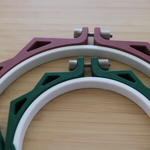 May include: Two circular embroidery hoops nested within each other. The larger outer hoop is maroon with a white inner ring, and the smaller inner hoop is dark green with a white inner ring. Both feature metal tightening screws at the top.