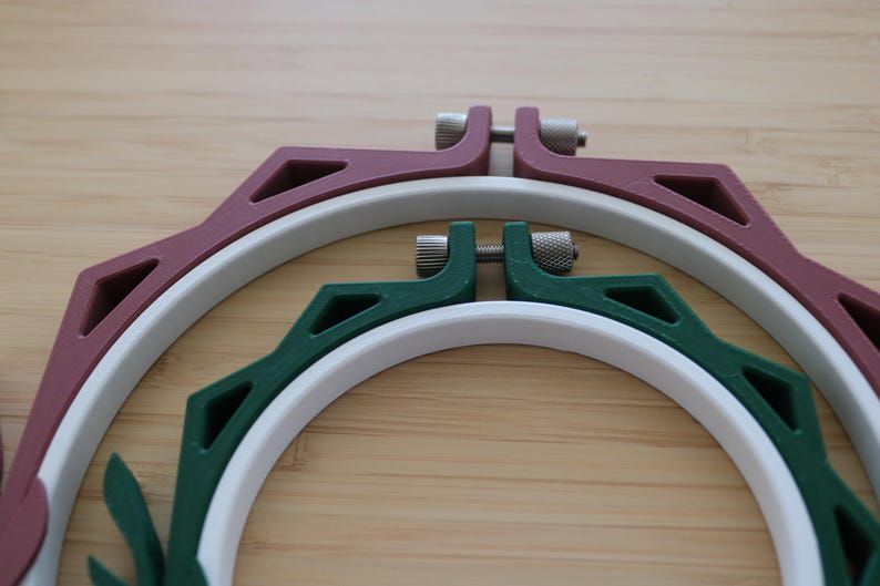 May include: Two circular embroidery hoops nested within each other. The larger outer hoop is maroon with a white inner ring, and the smaller inner hoop is dark green with a white inner ring. Both feature metal tightening screws at the top.