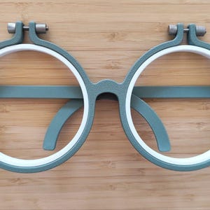 May include: A pair of circular embroidery hoops shaped like eyeglasses, featuring a textured sage green outer frame and a smooth white inner ring. Each hoop includes a small metal tension screw at the top for tightening the fabric during needlework.