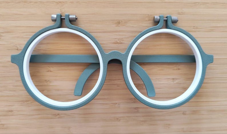 May include: A pair of circular embroidery hoops shaped like eyeglasses, featuring a textured sage green outer frame and a smooth white inner ring. Each hoop includes a small metal tension screw at the top for tightening the fabric during needlework.