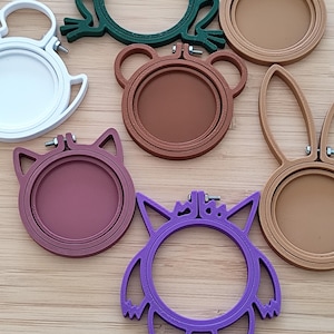 Puede incluir: Una colección de siete bastidores de bordado circulares en varios colores y formas de animales. El conjunto incluye un bastidor blanco, una rana verde oscuro, dos bastidores beige con orejas de conejo, un oso marrón, un gato rosa y uno morado.