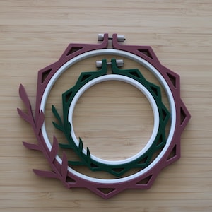 May include: Two nested embroidery hoops featuring a decorative outer frame with a leaf motif. The larger outer hoop is maroon, while the smaller inner hoop is dark green. Both hoops include a white circular inner ring and a metal tightening screw.