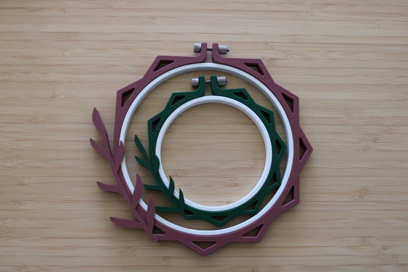 May include: Two nested embroidery hoops featuring a decorative outer frame with a leaf motif. The larger outer hoop is maroon, while the smaller inner hoop is dark green. Both hoops include a white circular inner ring and a metal tightening screw.