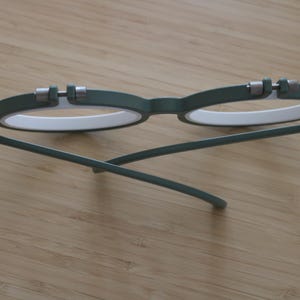 May include: A pair of round eyeglasses featuring a matte olive green frame with white inner rims. The glasses have silver-toned metal hinges at the top of each lens rim and thin, matching olive green temples that are folded inward.