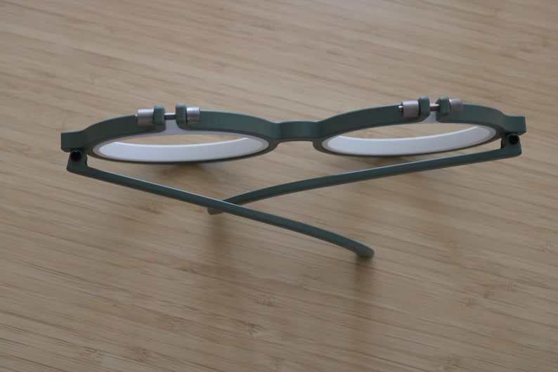 May include: A pair of round eyeglasses featuring a matte olive green frame with white inner rims. The glasses have silver-toned metal hinges at the top of each lens rim and thin, matching olive green temples that are folded inward.