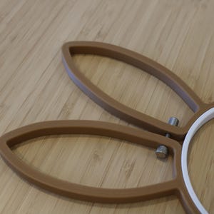 May include: A brown embroidery hoop featuring two elongated, ear-shaped extensions at the top. The frame is constructed from a solid brown material with a white inner ring and a small, metallic silver tension screw mechanism for tightening.