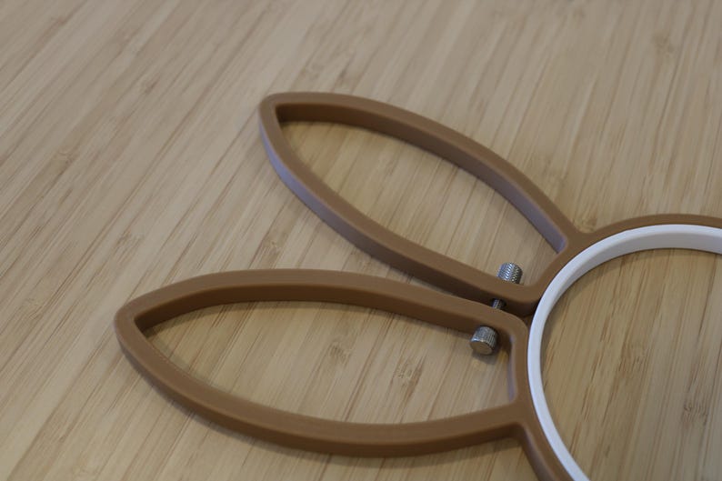 May include: A brown embroidery hoop featuring two elongated, ear-shaped extensions at the top. The frame is constructed from a solid brown material with a white inner ring and a small, metallic silver tension screw mechanism for tightening.