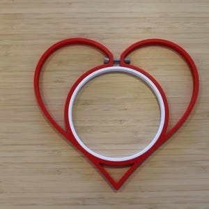 May include: A red heart-shaped embroidery hoop frame with a white circular inner hoop. The red frame features a metal tension screw at the top center, designed to hold fabric securely within the circular white insert for needlework projects.