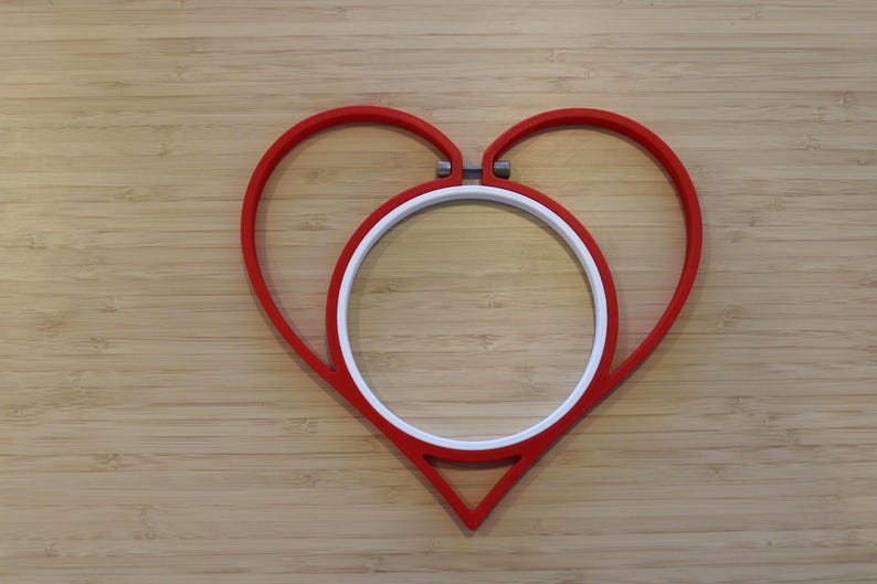 May include: A red heart-shaped embroidery hoop frame with a white circular inner hoop. The red frame features a metal tension screw at the top center, designed to hold fabric securely within the circular white insert for needlework projects.