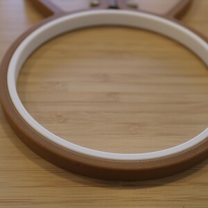 May include: A circular embroidery hoop featuring a brown outer ring and a white inner ring. The hoop is constructed from two distinct components that fit together to secure fabric during the embroidery process, showing a smooth finish.