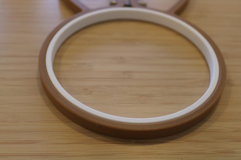 May include: A circular embroidery hoop featuring a brown outer ring and a white inner ring. The hoop is constructed from two distinct components that fit together to secure fabric during the embroidery process, showing a smooth finish.