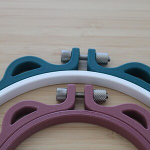 Op de afbeelding: Drie ronde borduurringen met een geschulpte buitenrand. De ringen zijn uitgevoerd in donkerblauwgroen, wit en oudroze. Elke ring is voorzien van een zilverkleurig metalen spanschroefmechanisme aan de bovenzijde om het frame vast te zetten.