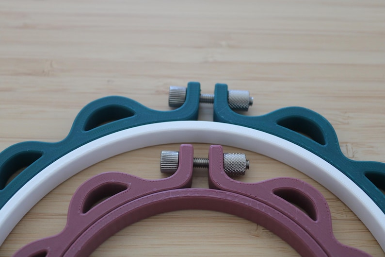 Op de afbeelding: Drie ronde borduurringen met een geschulpte buitenrand. De ringen zijn uitgevoerd in donkerblauwgroen, wit en oudroze. Elke ring is voorzien van een zilverkleurig metalen spanschroefmechanisme aan de bovenzijde om het frame vast te zetten.