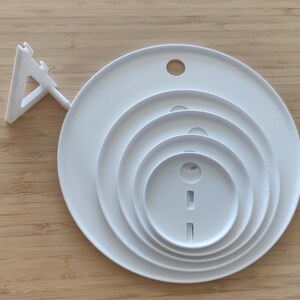May include: A white circular object featuring four concentric rings of varying sizes, each with a small circular or rectangular cutout. A triangular support stand is attached to the left side of the main circular base, allowing the object to be positioned upright.