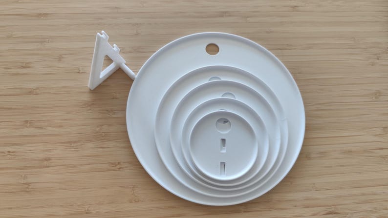 May include: A white circular object featuring four concentric rings of varying sizes, each with a small circular or rectangular cutout. A triangular support stand is attached to the left side of the main circular base, allowing the object to be positioned upright.