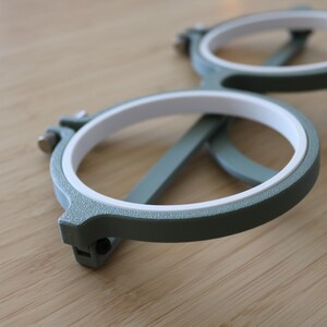 May include: A pair of circular embroidery hoops featuring a textured sage green outer frame and a smooth white inner ring. Each hoop includes a small metal tightening screw mechanism on the side to secure fabric in place during needlework projects.