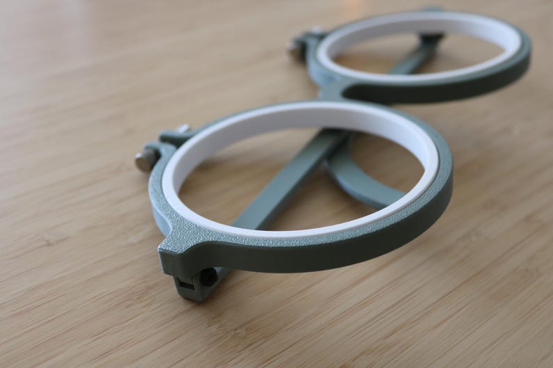 May include: A pair of circular embroidery hoops featuring a textured sage green outer frame and a smooth white inner ring. Each hoop includes a small metal tightening screw mechanism on the side to secure fabric in place during needlework projects.