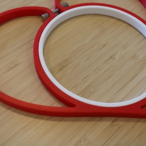 May include: A heart-shaped embroidery hoop featuring a vibrant red outer frame and a smooth white inner ring. The two components are connected by a small metal tension screw at the top, designed to hold fabric securely in place for needlework projects.