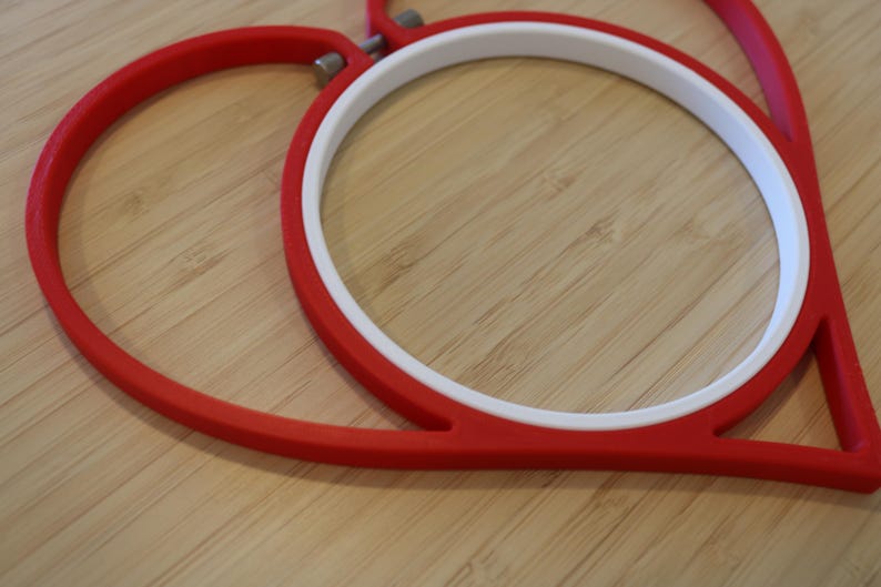 May include: A heart-shaped embroidery hoop featuring a vibrant red outer frame and a smooth white inner ring. The two components are connected by a small metal tension screw at the top, designed to hold fabric securely in place for needlework projects.