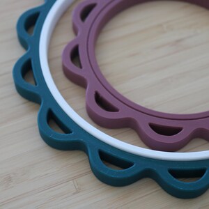 Op de afbeelding: Drie ronde onderzetters met geschulpte randen die in elkaar zijn genest. De buitenste ring is donkerblauwgroen, de middelste ring is wit en de binnenste ring is gedempt mauve. Elke onderzetter is voorzien van uitsparingen langs de rand.