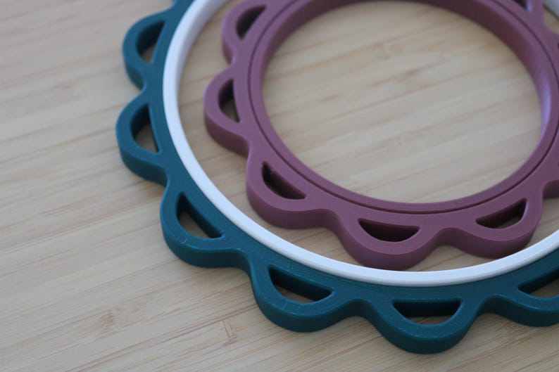 Op de afbeelding: Drie ronde onderzetters met geschulpte randen die in elkaar zijn genest. De buitenste ring is donkerblauwgroen, de middelste ring is wit en de binnenste ring is gedempt mauve. Elke onderzetter is voorzien van uitsparingen langs de rand.