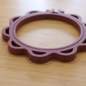 Op de afbeelding: Een cirkelvormige borduurring in een diepe kastanjebruine kleur met een geschulpte buitenrand en uitgesneden details. Het frame bestaat uit twee in elkaar grijpende ringen met een klein metalen schroefmechanisme aan de bovenkant.