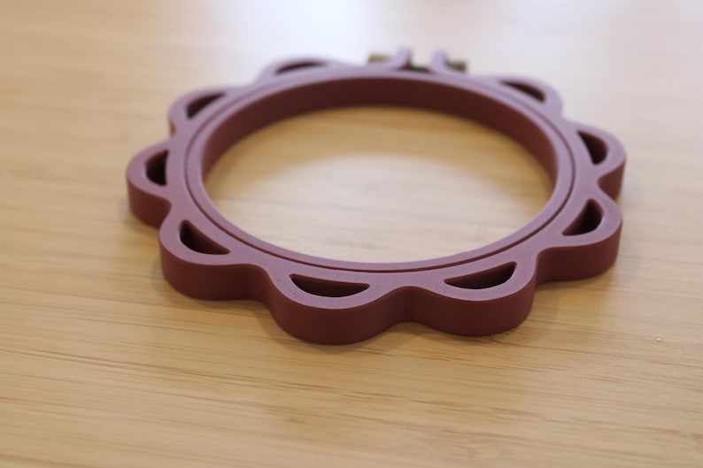 Op de afbeelding: Een cirkelvormige borduurring in een diepe kastanjebruine kleur met een geschulpte buitenrand en uitgesneden details. Het frame bestaat uit twee in elkaar grijpende ringen met een klein metalen schroefmechanisme aan de bovenkant.