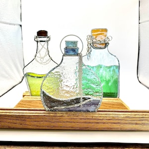 May include: Three stained glass bottles with different colored liquids inside. The bottles are on a wooden stand. The bottles are decorated with silver chains.