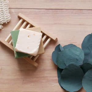 May include: Three square soaps in shades of green, beige, and cream rest on a wooden soap dish. A natural fiber washcloth and eucalyptus leaves are arranged on a light wood surface, creating a spa-like aesthetic. The soaps appear handmade.