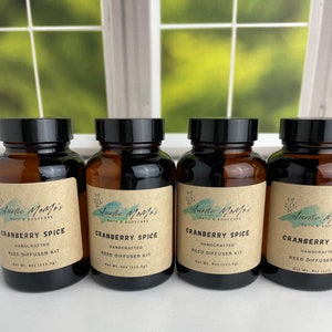May include: Four amber glass bottles with black lids, labeled "Cranberry Spice Reed Diffuser Kit." Each label features the text "Auntie Momo's Bath & Bodycare." The bottles are arranged in a row, with a window in the background.