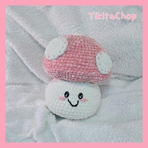 May include: A handmade crochet mushroom toy with a pink cap and white spots, atop a white base with a smiling face. The toy is set against a soft, white background, with the text "Tikitachop" in the top right corner.
