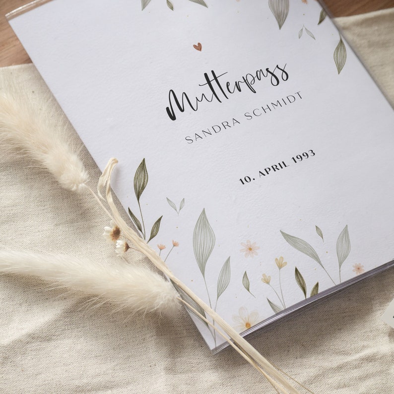 May include: A white book with the title "Mutterpass" and the name "Sandra Schmidt" printed on the cover. The date "10. April 1993" is also printed on the cover. The book is decorated with a floral design in shades of green and beige.