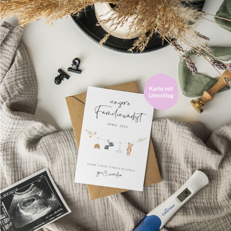 May include: A white card with a brown envelope. The card has a cute illustration of a clothesline with baby items hanging on it. The text on the card reads "Unsere Familie w&auml;chst" and "April 2024". The text on the envelope reads "Karte mit Umschlag".
