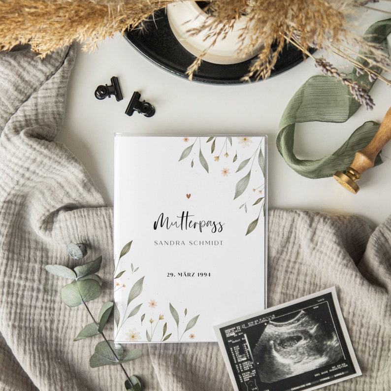 May include: A white book with a floral design and the text "Mutterpass Sandra Schmidt 29. M&auml;rz 1994".