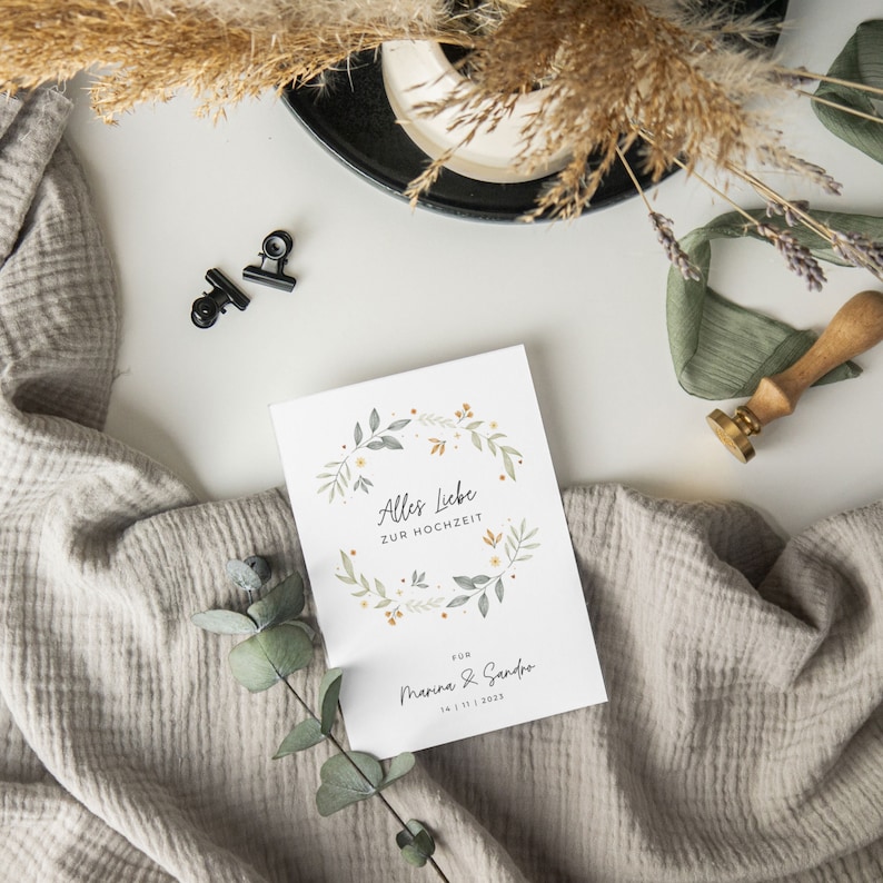 May include: A white card with a floral wreath design and the text "Alles Liebe zur Hochzeit" for Marina & Sandro, 24.11.2023. The card is on a white surface with a brown and white blanket, a small bouquet of dried flowers, and a black paper clip.