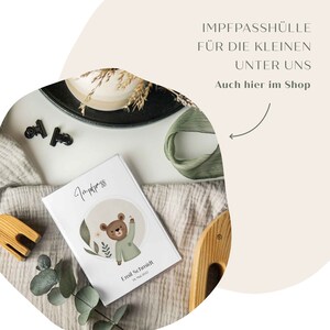 May include: A white card with a green and brown bear illustration and the text "Impfpass" and "Emil Schmidt 14. Mai 2022". The card is on a light brown blanket with a green ribbon and a wooden tray.