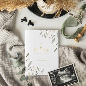 May include: A white book with a floral design and gold lettering. The book says "Mutterpass" and "Sandra Schmidt" with the date "26. M&auml;rz 1994".