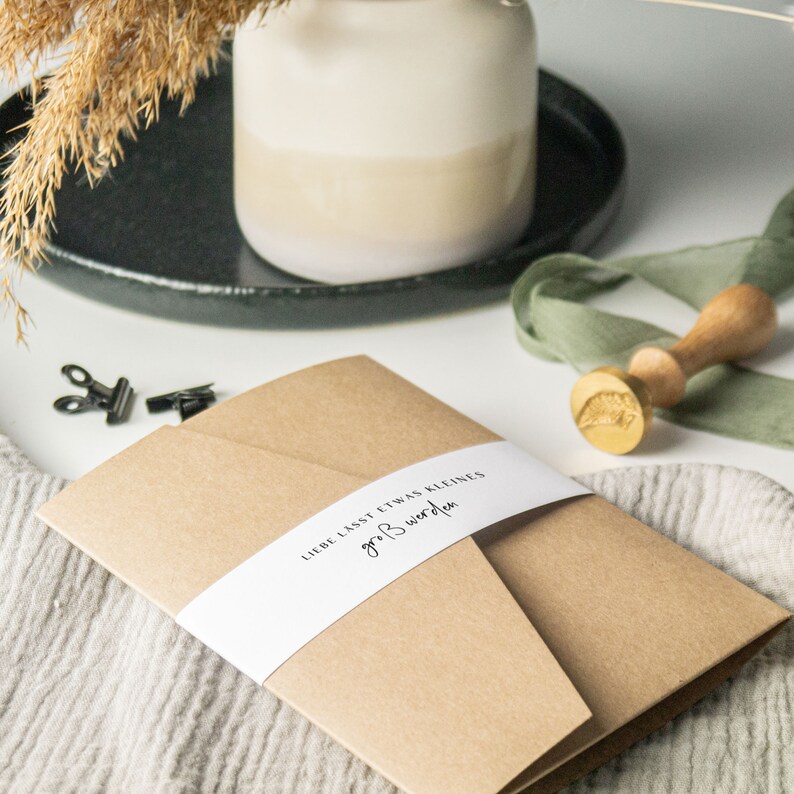 May include: A brown paper envelope with a white label that reads "Liebe lasst etwas kleines gro&szlig; werden". The envelope is sitting on a white surface with a green ribbon and a gold wax seal.