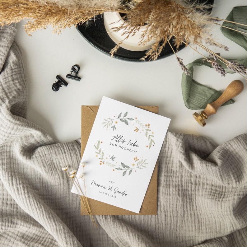 May include: A white card with a floral wreath design and the text "Alles Liebe zur Hochzeit" for "Marina & Sandro" with the date "14.11.2023".