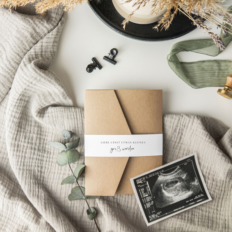 May include: A brown envelope with a white label that reads "Liebe lasst etwas kleines gro&szlig; werden". The envelope is on a white surface with a piece of fabric and a eucalyptus branch. There is also a black and white ultrasound image.