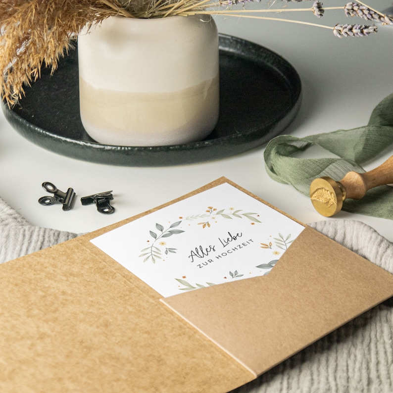 May include: A brown envelope with a pocket on the front. The pocket contains a white card with a floral wreath and the text "Alles Liebe zur Hochzeit".